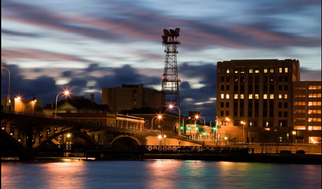 Cityscape at dusk with illuminated buildings and a bridge over water.