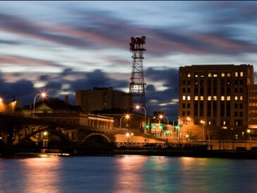 Cityscape at dusk with illuminated buildings and a bridge over water.