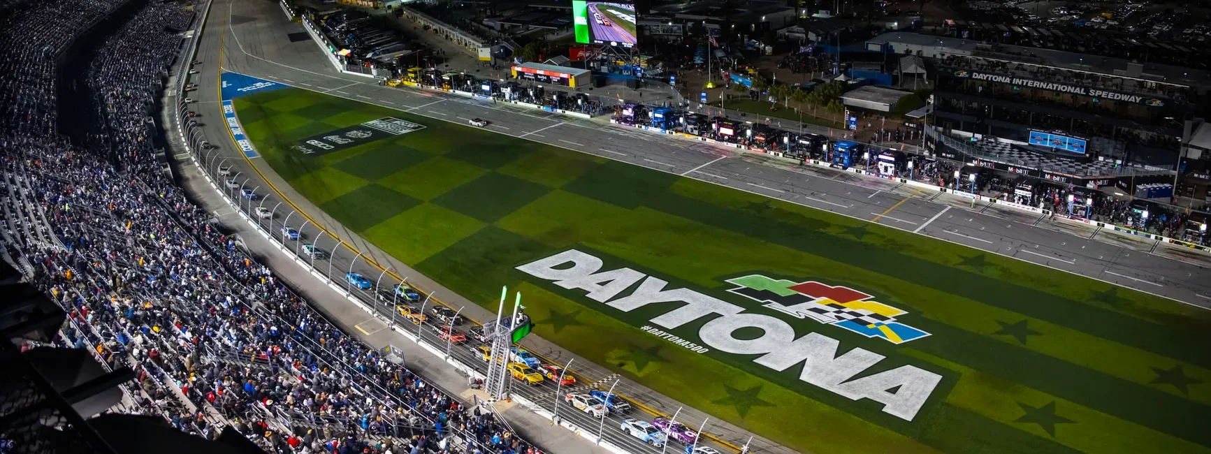 Nighttime view of Daytona International Speedway with race cars on track.
