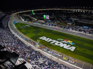 Nighttime view of Daytona International Speedway with race cars on track.