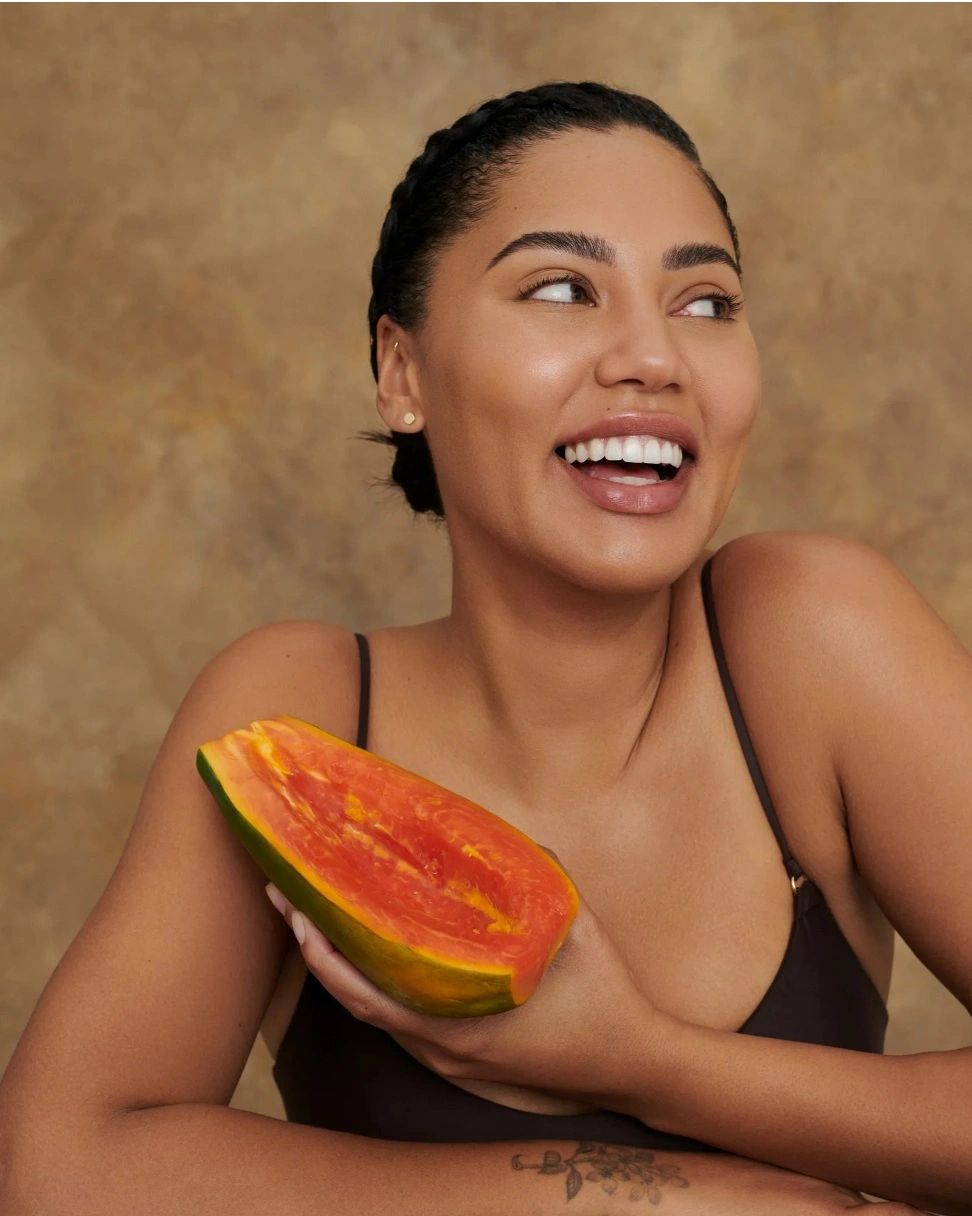 Smiling woman holding a ripe papaya against a warm brown background.