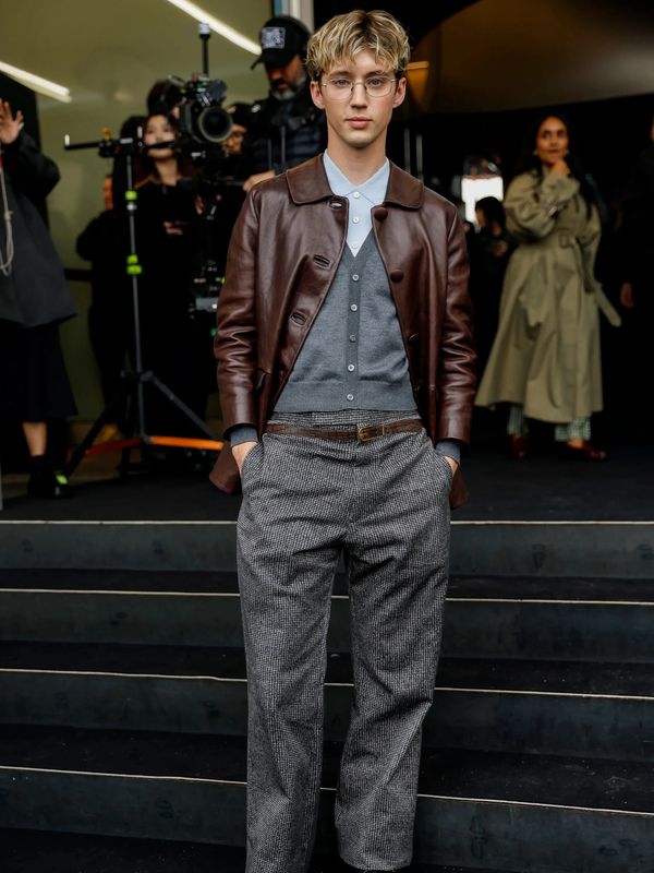 Man in brown leather jacket and gray pants standing with hands in pockets.